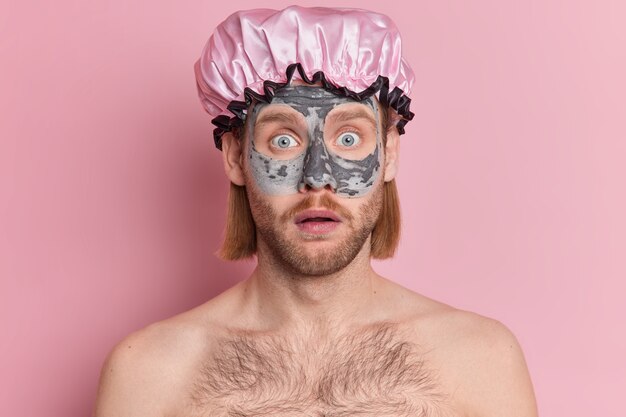 Portrait d'homme barbu surpris regarde choqué porte un chapeau de douche applique un masque d'argile nourrissant pour le rajeunissement et la réduction des rides se tient avec les épaules nues.