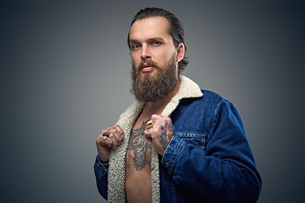 Photo gratuite portrait d'homme barbu avec des tatouages sur les bras et la poitrine portant une veste en jean sur fond gris.