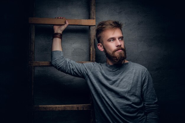 Portrait d'un homme barbu urbain posant près de la vieille échelle vintage sur fond gris.