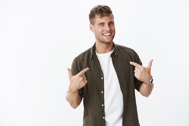 Portrait d'un homme blond charismatique, satisfait de lui-même et fier, pointant sur lui-même indiquant le corps et souriant largement comme voulant être choisi sur un mur gris