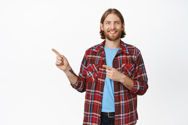 Portrait d'un homme blond souriant avec barbe, pointant les doigts vers la gauche et ayant l'air heureux, portant des vêtements décontractés, montrant une bannière de vente à prix réduit, debout sur fond blanc