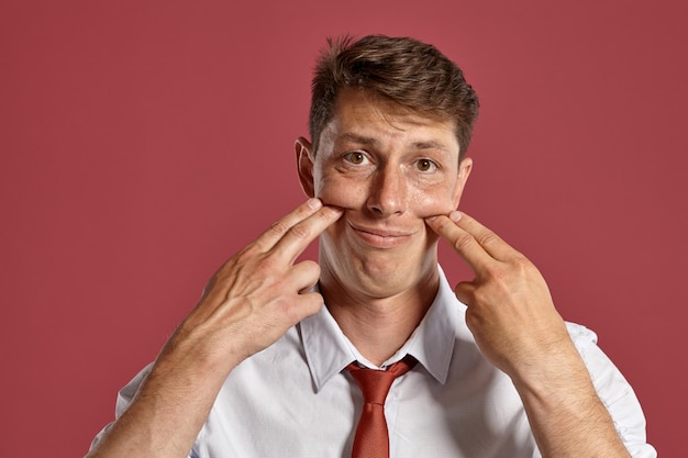 Portrait D'un Homme Brun Comique Aux Yeux Bruns, Vêtu D'une Chemise Blanche Et D'une Cravate En Terre Cuite. Il S'amuse En Posant Dans Un Studio Sur Fond Rouge. Concept De Gesticulation Et Si