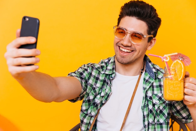 Portrait d'homme brun à lunettes de soleil orange tenant un verre à cocktail et prenant selfie sur l'espace orange.