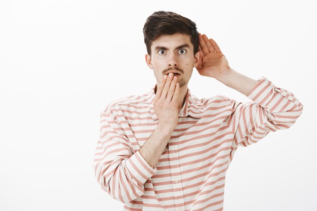 Portrait d'un homme caucasien séduisant intrigué choqué en chemise rayée à la mode, tenant la main près de l'oreille et de la bouche, entendant une conversation ou écoutant, entendre quelque chose de choquant et d'intéressant