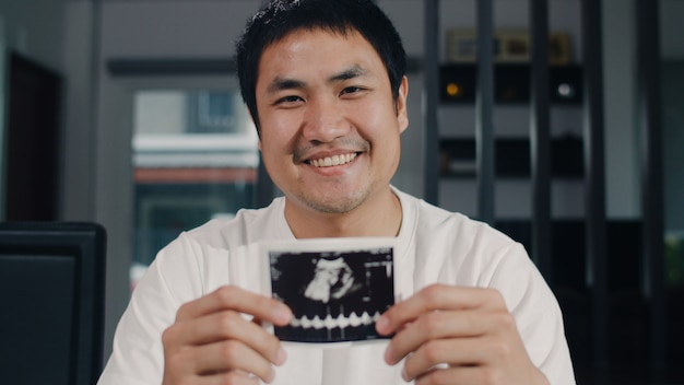 Portrait homme chinois asiatique montrent et à la recherche de bébé photo ultrasons dans sa femme à la maison. Jeune homme détendu sourire toothy à la recherche à la caméra assis sur la table dans le salon à la maison le matin.