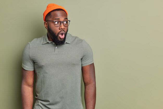 Portrait d'un homme choqué regarde avec les yeux et la bouche largement ouverts réagit émotionnellement sur des nouvelles étonnantes porte un t-shirt et des lunettes csual chapeau orange isolé sur mur vert