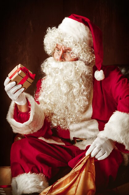 Portrait d'homme en costume de père Noël - avec une barbe blanche luxueuse, un chapeau du père Noël et un costume rouge au studio rouge assis avec des cadeaux