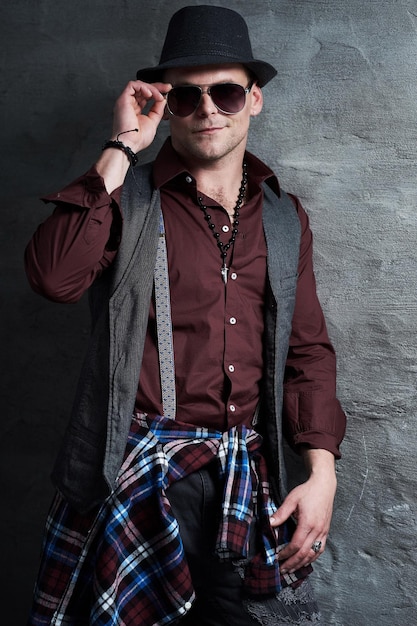 Portrait d'un homme décontracté vêtu d'une chemise, d'un gilet et de lunettes de soleil sur fond gris.