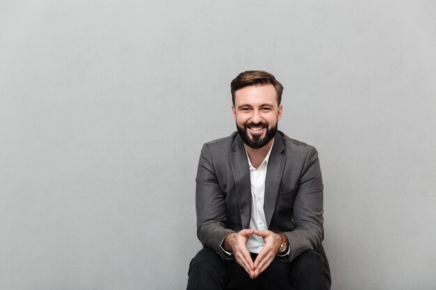 Portrait de l'homme détendu reposant assis sur une chaise dans le bureau et souriant à la caméra mettant les mains ensemble, isolé sur gris