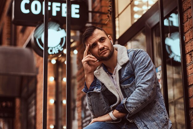 Portrait d'un homme élégant pensif portant une veste en jean avec des écouteurs sans fil tenant du café à emporter à l'extérieur du café.