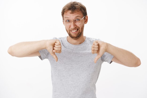 Portrait d'un homme européen mignon et drôle mécontent à lunettes et t-shirt gris montrant les pouces vers le bas et grimaçant de ne pas aimer exprimer sa désapprobation d'être insatisfait