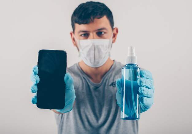 Portrait d'un homme à fond clair tenant un téléphone et un désinfectant pour les mains dans des gants médicaux et un masque