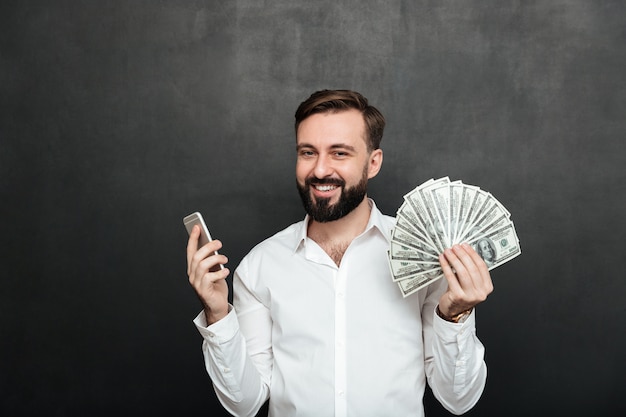 Portrait d'homme gai en chemise blanche remportant beaucoup d'argent dollar en utilisant son smartphone, être joyeux sur gris foncé