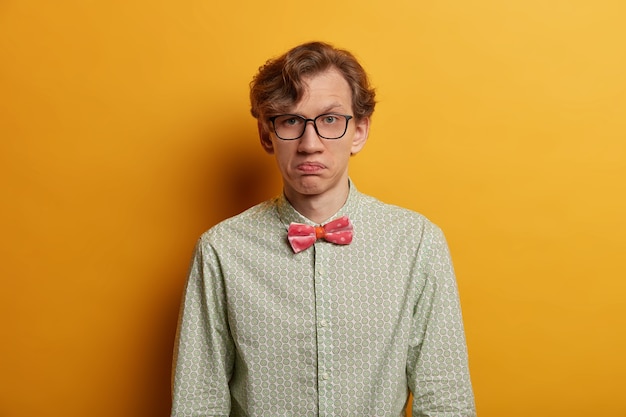 Photo gratuite portrait d'un homme hésitant sérieux et non impressionné, porte des lèvres, regarde dubitatif, porte des lunettes optiques et une chemise formelle, a les cheveux ondulés, des modèles à l'intérieur sur un mur jaune. expressions de visage humain