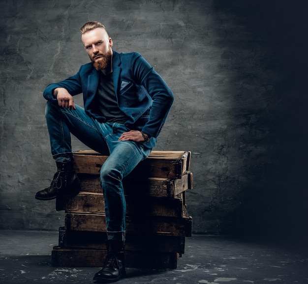 Portrait d'homme hipster barbu rousse vêtu d'une veste bleue posant sur une boîte en bois sur fond gris.