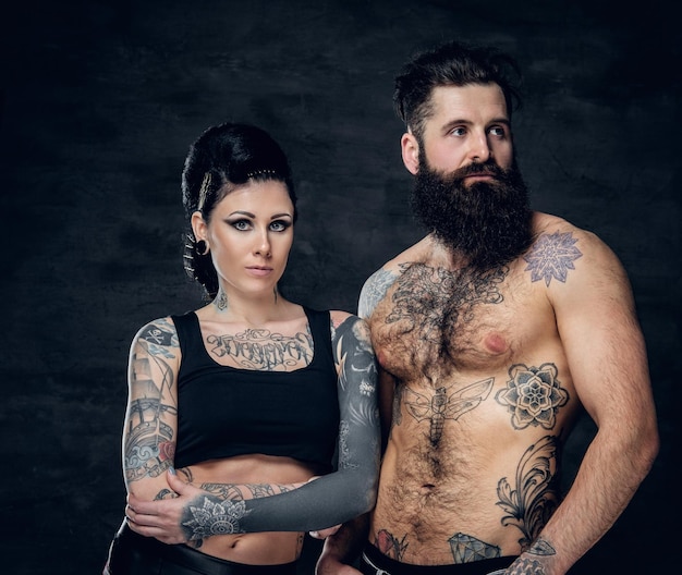 Portrait d'homme hipster barbu tatoué et torse nu et d'une femme brune avec de l'encre de tatouage sur son torse sur fond gris foncé.