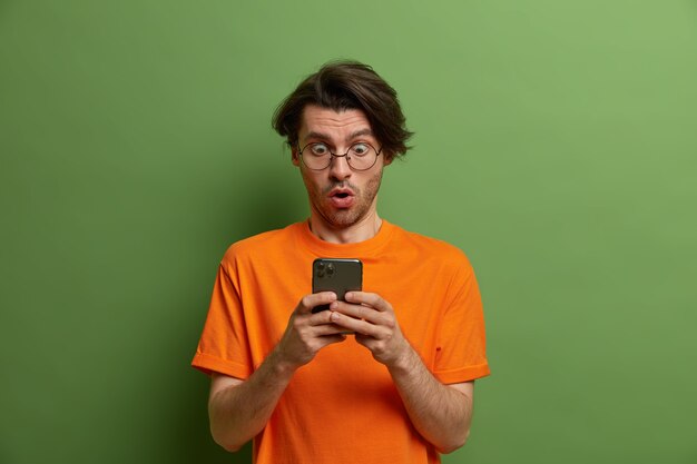 Portrait d'un homme impressionné surpris regarde l'écran du smartphone, ne peut pas en croire ses propres yeux, reçoit un message choquant, ouvre la bouche et retient son souffle, porte un t-shirt orange, pose contre le mur vert