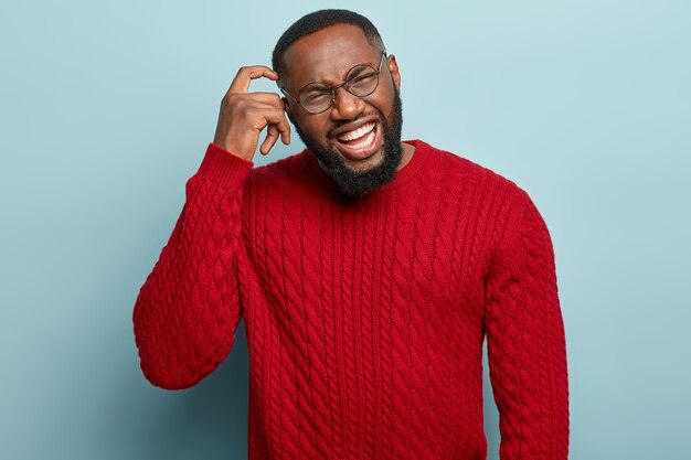 Portrait d'un homme mécontent à la peau sombre se gratte la tête, a une expression insatisfaite, fronce les sourcils et montre des dents blanches, porte un pull rouge, isolé sur un mur bleu. Concept de sentiments négatifs