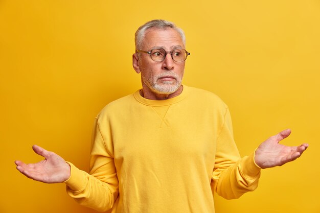 Portrait d'homme mûr barbu hésitant perplexe étend les paumes et les visages choix difficile semble incertain porte des lunettes optiques cavalier occasionnel a confondu l'expression isolée sur mur jaune