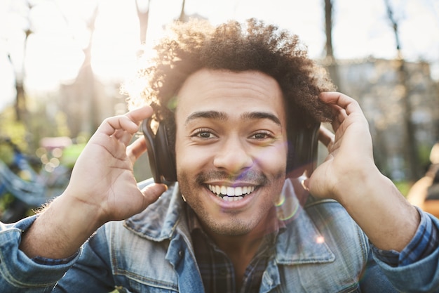 Portrait d'un homme à la peau sombre adulte positif souriant largement alors qu'il était assis dans le parc, écoutant de la musique dans les écouteurs et les tenant avec les mains pour mieux entendre.