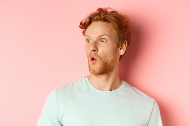 Portrait d'un homme roux surpris avec une barbe, regardant à gauche et disant wow, levant les sourcils étonnés, debout sur fond rose.