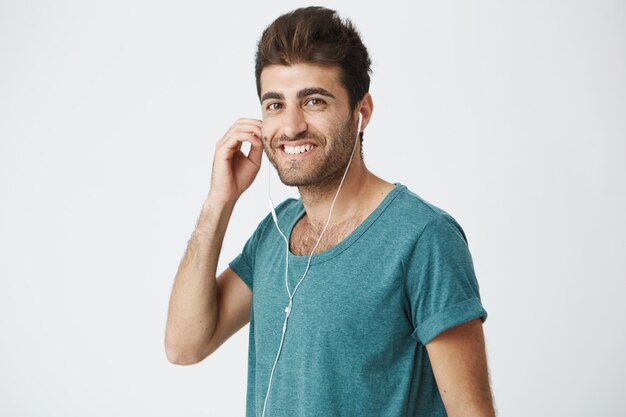 Portrait d'un homme séduisant, beau et élégant, souriant portant des écouteurs blancs. Écoute du nouvel album de son groupe préféré en utilisant l'application de musique en ligne