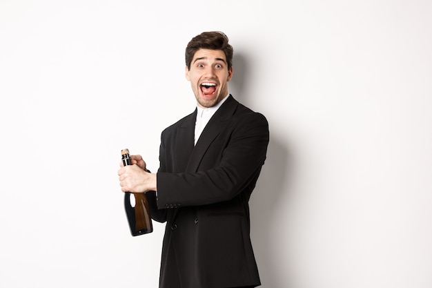 Portrait d'un homme séduisant en costume noir, faisant un clin d'œil à la caméra et ouvrant une bouteille de champagne, célébrant le nouvel an, debout sur fond blanc.