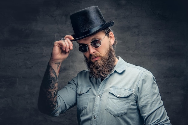 Portrait d'un homme sérieux et barbu portant des lunettes de soleil et un chapeau cylindrique.