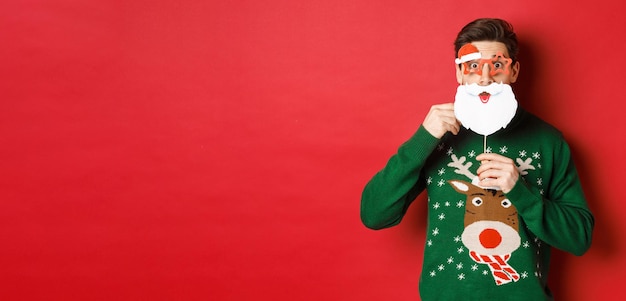 Portrait d'un homme surpris en pull de noël vert tenant un masque drôle de père noël célébrant le nouveau ...