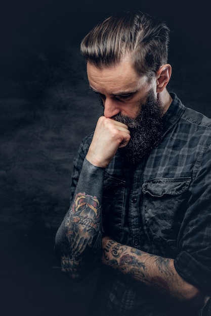 Le portrait d'un homme tatoué et barbu en chemise touche sa tête sur un fond sombre.