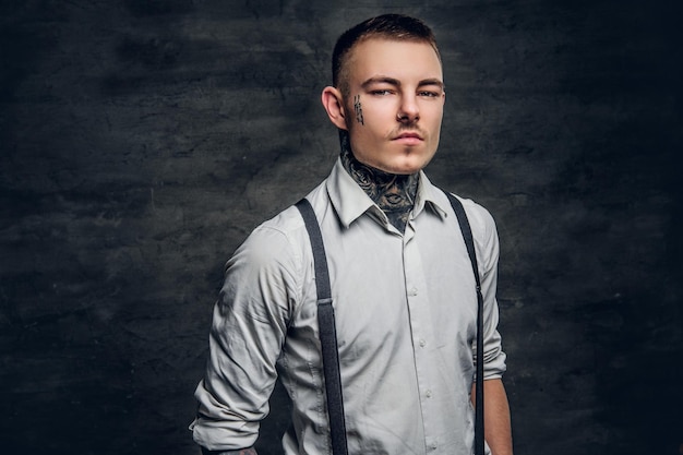 Portrait d'un homme tatoué sur le visage et les bras, vêtu d'une chemise blanche sur fond gris.