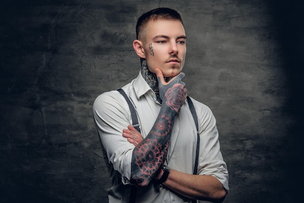 Portrait d'un homme tatoué sur le visage et les bras, vêtu d'une chemise blanche sur fond gris.