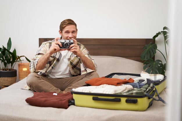 Portrait d'un homme touriste assis avec une valise sur le lit tient l'appareil photo un sac de valise avec des vêtements continue