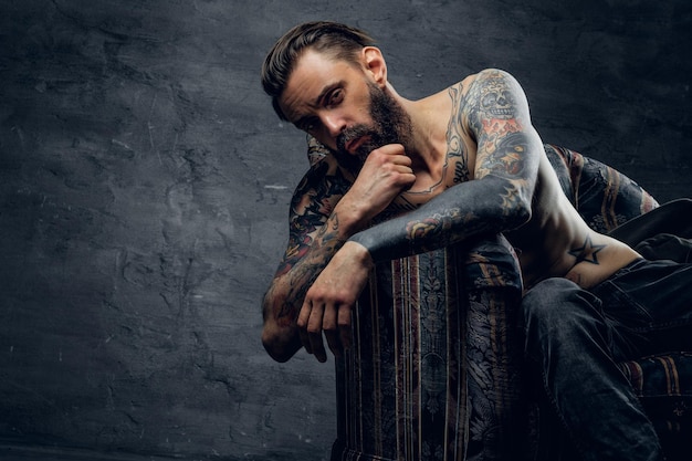 Portrait d'un homme triste tatoué désespéré, qui est assis sur un fauteuil dans une cachette sombre.