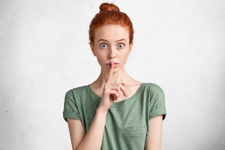 Photo gratuit portrait horizontal de femme aux taches de rousseur choqué avec noeud de cheveux gingembre, montre signe de silence, demande de garder les informations confidentielles ou privées, pose contre blanc