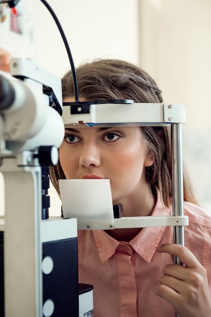 Portrait horizontal de femme européenne ciblée testant la vue tout en regardant à travers le microbioscope, assis dans un bureau spécialisé, voulant choisir les lunettes appropriées pour mieux voir