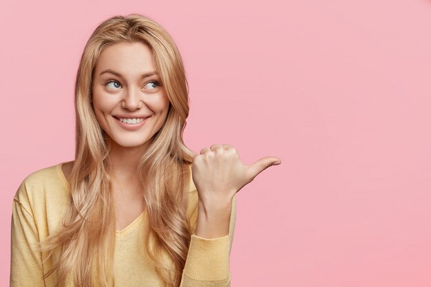 Portrait horizontal de femme heureuse aux cheveux blonds et aux yeux bleus, shing smile, fait la publicité d'un produit, indique de côté, pose contre le mur rose avec espace de copie pour votre texte publicitaire