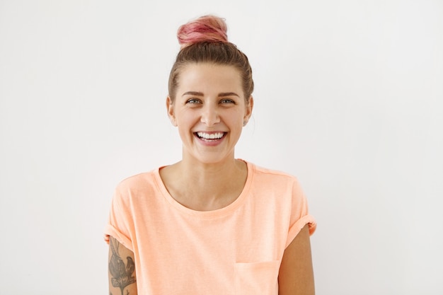 Portrait horizontal de femme joyeuse avec sourire attrayant, ayant chignon et tatouage rose, portant des vêtements décontractés,