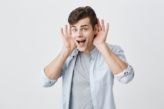 Portrait horizontal d'un jeune homme caucasien surpris, étonné par les gros prix de vente, a un look choqué, les yeux sortis et la mâchoire baissée, les paumes ouvertes derrière les oreilles.