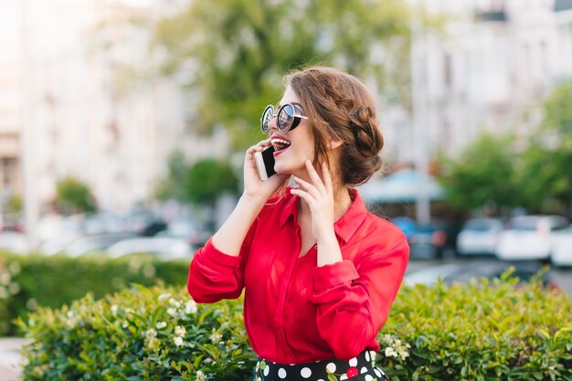 Portrait horizontal de jolie fille à lunettes de soleil marchant dans le parc. Elle porte un chemisier rouge et une belle coiffure. Elle parle au téléphone et sourit au loin.