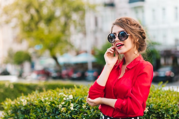 Portrait horizontal de jolie fille à lunettes de soleil posant à la caméra dans le parc. Elle porte un chemisier rouge et une belle coiffure. Elle regarde au loin.