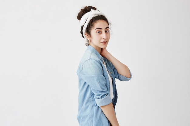 Portrait horizontal de modèle féminin positif garde la main sur le cou, avec des cheveux foncés et ondulés en chignon, regarde rêveusement, se réjouit d'avoir des vacances, pose contre un mur blanc avec copie espace