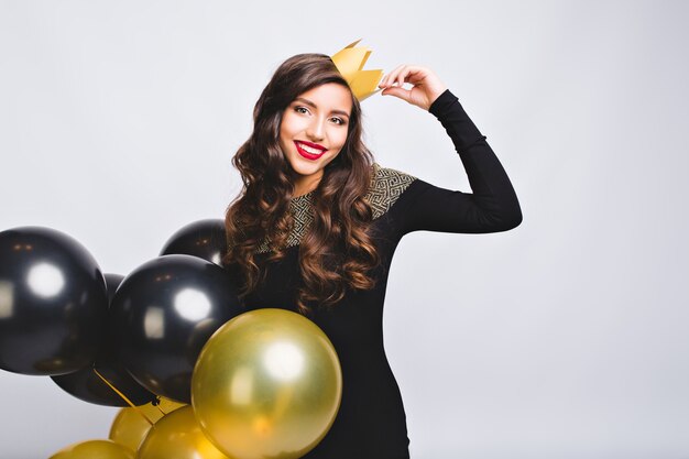 Portrait incroyable jolie femme célébrant les vacances, tenant des ballons or et noirs, vêtue d'une robe noire et couronne jaune, s'amuser, partie