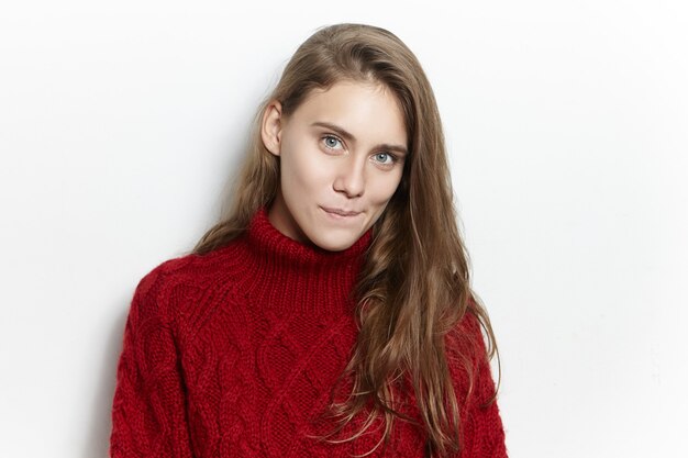 Portrait d'incroyable magnifique jeune femme européenne avec une longue coiffure lâche posant isolé dans un pull marsala confortable, regardant avec une expression faciale curieuse mystérieuse, mordre les lèvres