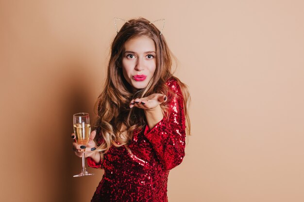 Portrait intérieur d'adorable femme européenne en robe rouge envoi de baiser d'air et tenant un verre de champagne