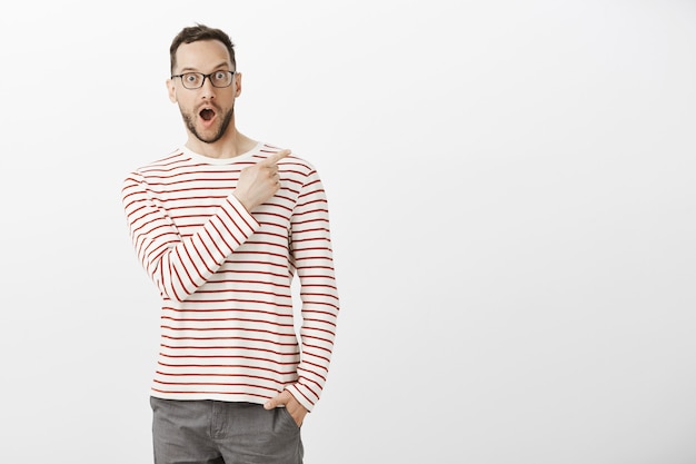 Portrait Intérieur D'un Bel Homme Heureux Impressionné Dans Les Lunettes, Pointant Vers Le Coin Supérieur Droit, Laissant Tomber La Mâchoire D'émotions Positives