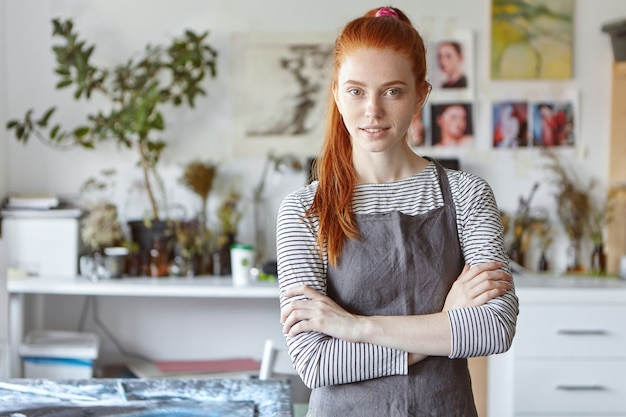 Portrait intérieur de charmante jeune femme artisanale rousse confiante portant un tablier gris debout dans son atelier, gardant les mains croisées et souriant, prêt pour le processus de création et de travail acharné