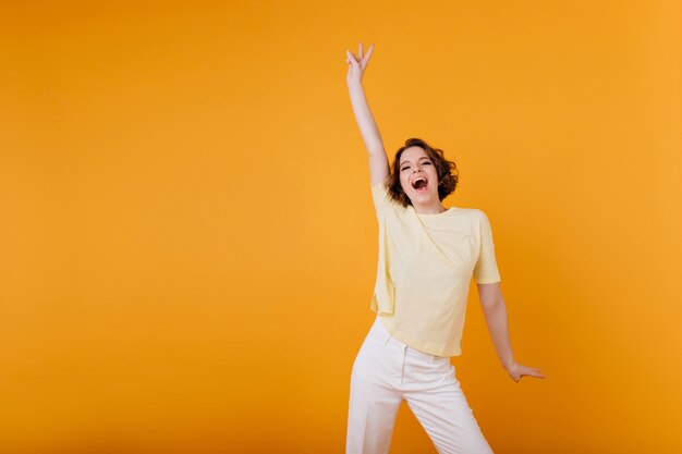 Portrait intérieur de femme pâle aux cheveux noirs debout avec la main vers le haut. fille brune raffinée en t-shirt jaune détendu pendant la séance photo sur un mur lumineux.