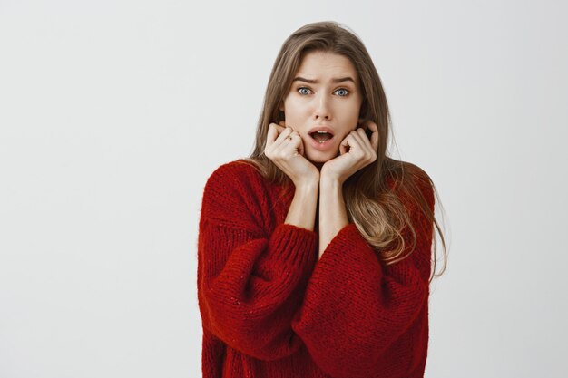 Portrait intérieur de femme timide européenne inquiète en pull rouge lâche, se tenant la main sur le menton et la mâchoire tombante, étant anxieux et nerveux, sympathisant un ami à l'hôpital sur un mur gris