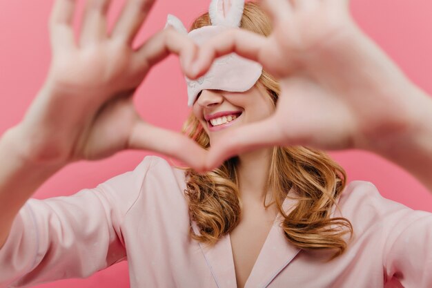 Portrait intérieur d'une fille blonde romantique en masque pour les yeux posant avec signe d'amour. Jolie jeune femme en masque de sommeil et pyjama s'amusant dans sa chambre.